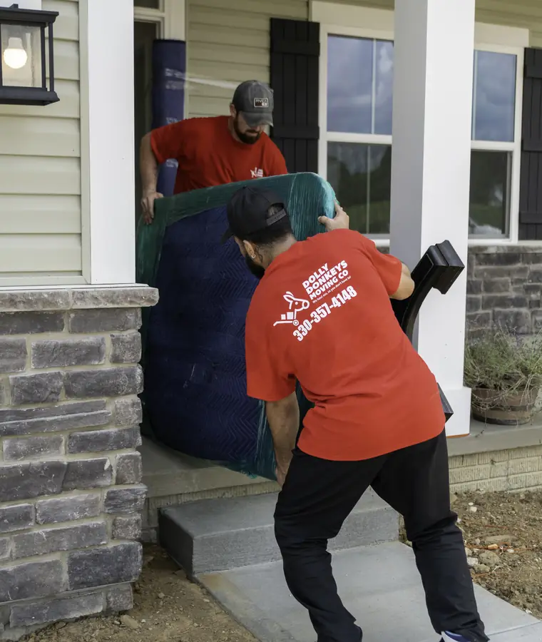 Two Dolly Donkeys movers carrying a blanket-wrapped piece up to the porch